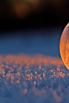 Close-up shot of a frozen bubble with warm reflections resting on a snowy surface at twilight.